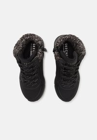 Scarpe nere alte con un colletto testurizzato e peloso, caratterizzate da lacci neri, accenti metallici e una punta forata per la ventilazione.