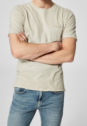 Hellgraues T-Shirt mit Rundhalsausschnitt, kurzen Ärmeln und einer linken Brusttasche, kombiniert mit taillierten blauen Jeans.