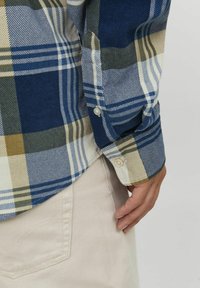Camisa a cuadros con colores azul, verde y beige, con textura de pana, puños de botón y dobladillo redondeado.