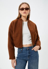 Giacca in pile marrone con zip, dal design corto e colletto alto, abbinata a una camicia bianca e jeans a vita alta blu.