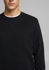 Sudadera negra con cuello redondo, mangas largas y una superficie texturizada. Presenta un detalle de costura en el escote y un ajuste holgado.