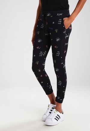 Person trägt schwarze Jogginghosen mit bunten Kritzeleien und motivierenden Texten, kombiniert mit weißen Adidas-Sneakern mit schwarzen Streifen.