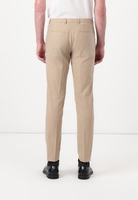 Pantaloni beige sartoriali con vestibilità slim, dotati di due tasche posteriori, passanti per cintura e una texture di tessuto liscia. Indossati con scarpe eleganti nere.