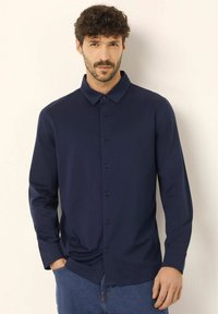 Camicia blu navy a maniche lunghe con colletto a punta, realizzata in tessuto morbido, con sei bottoni sul davanti e una leggera lucentezza.