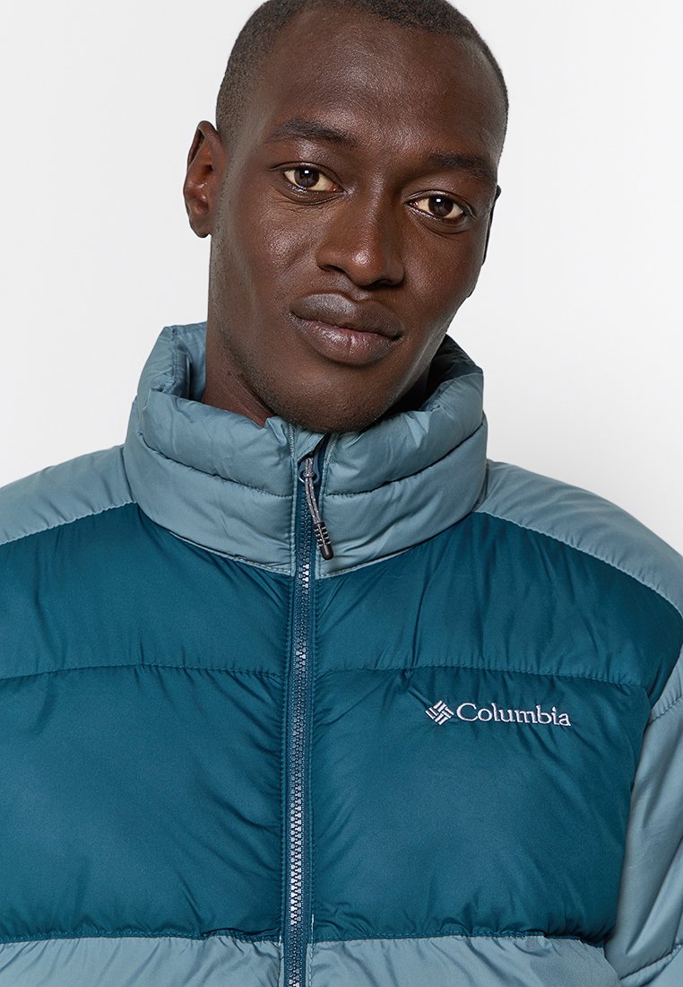 Giacca isolante color verde cenere e azzurro chiaro con colletto alto, dotata di zip frontale e logo Columbia sul petto. Design a collo d'oca strutturato.