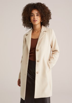 Manteau beige en tissu doux, avec un col à revers, des manches longues et quatre grands boutons sur le devant. Couleur unie, design structuré.