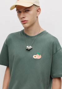 Grünes Baumwoll-T-Shirt mit einem gestickten Pfirsichdesign. Trägt eine silberne Halskette und eine hellfarbige Kappe.