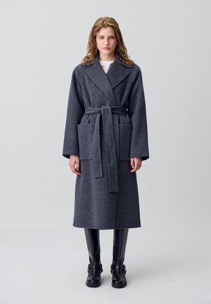 Manteau long en laine grise avec une ceinture, grandes poches avant, revers larges et coupe droite. Porté avec des bottes noires montantes.