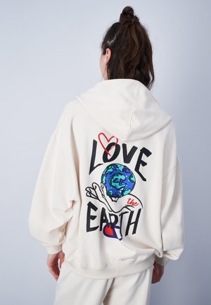 Persoon draagt een witte hoodie met een afbeelding van de aarde die door een hand wordt vastgehouden en de tekst "Love the Earth" op de achterkant.