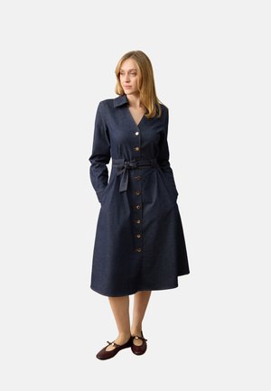 Femme portant une robe bleu marine boutonnée manches longues, ceinture à la taille et chaussures plates bordeaux, debout les mains dans les poches.