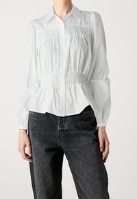 Weiße Bluse mit Knopfverschluss und Raffung an der Taille, langen Puffärmeln und einem taillierten Kragen. Kombiniert mit dunklen Jeans.