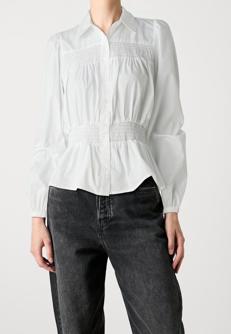 Weiße Bluse mit Knopfverschluss und Raffung an der Taille, langen Puffärmeln und einem taillierten Kragen. Kombiniert mit dunklen Jeans.