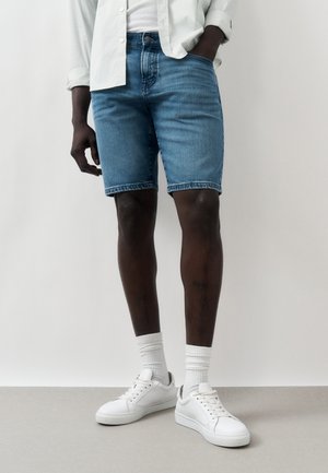 Személy világoskék farmer rövidnadrágot, fehér zoknit, fehér tornacipőt és fehér inget visel, az egyik kezével a zsebében áll egy szürke padlón.