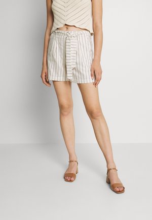Shorts rayés beige avec un nœud devant, fabriqués en tissu léger. Assortis à des sandales à talons blocs de couleur taupe, avec une bride autour de la cheville.