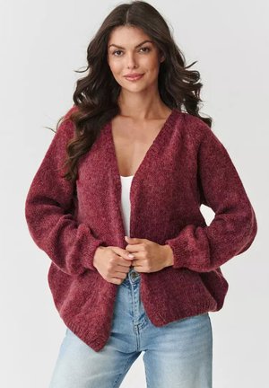 Burgundowy, oversize sweter z dekoltem w serek, długimi rękawami balonowymi i fakturowanym wzorem dzianiny, w połączeniu z białą bluzką i jasnymi dżinsami.