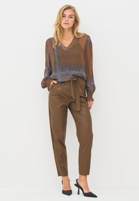 Blouse marron transparente à motif géométrique, associée à un pantalon taille haute en similicuir marron et des escarpins noirs pointus.
