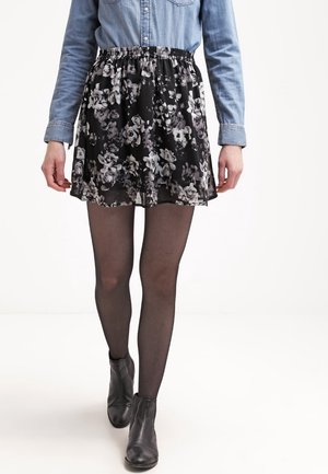Jupe florale noire en tissu transparent avec une taille élastique, associée à des collants noirs et des bottines, portée avec un haut ajusté.