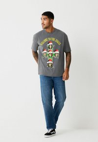 Szürke pamut póló, színes koponya grafikával, Mikulás sapkákkal. Kék farmerrel és fekete tornacipővel párosítva. Laza szabású és kialakítású.