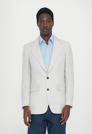 Lichtgrijze blazer met revers en twee voorzakken. Voorzien van zwarte knopen en gecombineerd met een lichtblauw shirt en donkere jeans.