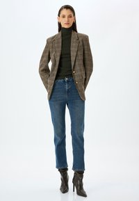 Blazer de cuadros en marrón y crema con botones dorados, suéter de cuello alto verde, vaqueros azules y botas de tacón marrones; texturas suaves y corte entallado.