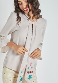 Blusa beige clara con lazo en el frente, mangas de encaje y bordado floral en rojo, rosa y verde. Tejido suave y ajuste relajado.