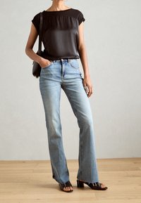 Kobieta w czarnej bluzce z krótkim rękawem, jasnoniebieskich dżinsach o wysokim stanie z surowymi krawędziami, czarnych butach na obcasie odkrytych z przodu i niesie czarną torbę na ramię.