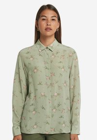 Lys grøn bluse med blomsterprint; lange ærmer, spidse krave, knaplukning foran og glat, let stof.