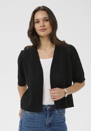 Kobieta ubrana w czarny kardigan z krótkim rękawem o otwartym splocie, nałożony na biały top i niebieskie dżinsy, stoi z jedną ręką na krawędzi kardiganu.