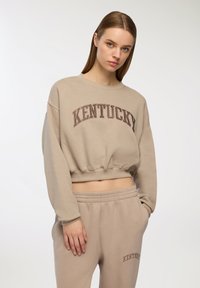 Felpa corta beige con "KENTUCKY" stampato in marrone ad arco. Tessuto morbido, maniche lunghe, orlo elastico. Abbinata a pantaloni della tuta coordinati.