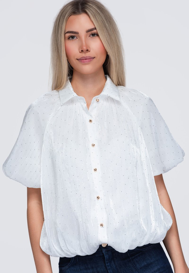 Femme aux longs cheveux blonds portant un chemisier blanc à manches courtes avec de petits pois et des boutons dorés, posant devant un fond uni.
