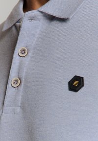 Lichtblauwe polo met een gestructureerde brei, drie knopen en een zwart zeshoekig logopatch met drie gele lijnen.