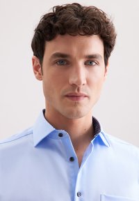 Jasnoniebieska koszula zapinana na guziki z kołnierzykiem, z wzorzystym ciemnym wykończeniem kołnierza i czarnymi guzikami. Gładki materiał i dopasowany krój.