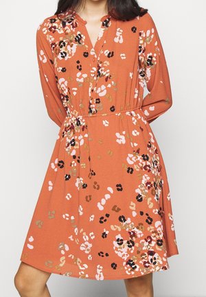 Frau trägt ein knielanges, langärmliges, orangenes Kleid mit einem mehrfarbigen abstrakten Blumenmuster und einem mit Knöpfen versehenen Ausschnitt.