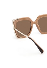 Hnědé oversized sluneční brýle s lesklým, osmiúhelníkovým hnědým rámečkem se zlatými detaily a černým ramínkem. Skla jsou tmavě hnědá.