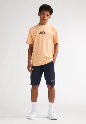 Camiseta de algodón color melocotón con gráfico de texto negro, combinada con pantalones cortos azul marino y zapatillas blancas. El modelo está de pie en posición neutral sobre un fondo claro.