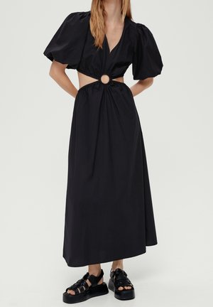 Kobieta w czarnej maxi sukience z bufkami na rękawach, bocznymi wycięciami, centralnym elementem obręczy oraz czarnych sandałach na platformie.