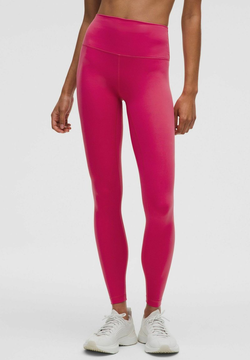 Leggings roses taille haute avec une texture lisse et extensible, présentant un design ajusté. Portés avec des chaussures de sport de couleur claire.