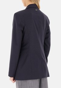 Blazer blu navy con una sottile texture a puntini, caratterizzato da un colletto classico e un piccolo logo del marchio sul retro. Vestibilità sartoriale con tasche laterali.