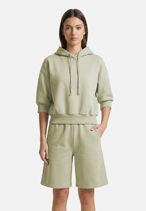 Jonge vrouw draagt een lichtgroene hoodie met 3/4 mouwen en bijpassende shorts tot op de knie, staand met één hand in de zak.