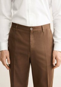 Pantaloni marroni su misura realizzati in tessuto morbido, con taglio dritto, chiusura a un bottone, tasche laterali e pieghe pronunciate.