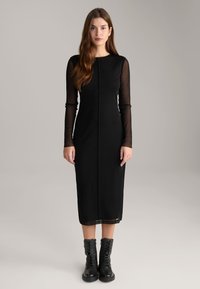 Schwarz-langärmeliges Kleid mit tailliertem Oberteil und halbdurchsichtigen Ärmeln, das einen geraden Saum hat. Getragen mit schwarzen Schnürkampfschuhen.