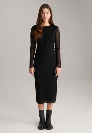Schwarz-langärmeliges Kleid mit tailliertem Oberteil und halbdurchsichtigen Ärmeln, das einen geraden Saum hat. Getragen mit schwarzen Schnürkampfschuhen.