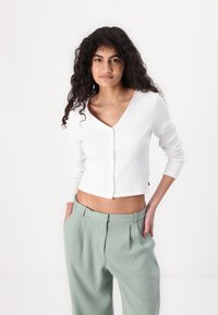 Cardigan blanc côtelé avec un col en V, fermeture par boutons et manches longues, associé à un pantalon vert clair taille haute.