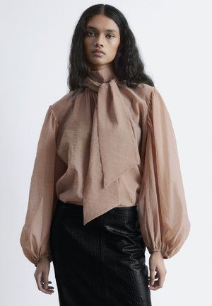 Pfirsichfarbene Bluse mit hohem Kragen und übergroßer Schleifendetail, mit puffigen Ärmeln; kombiniert mit einem strukturierten schwarzen Lederrock.