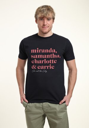 Joven de cabello claro con una camiseta negra que muestra en texto rosa los nombres "Miranda, Samantha, Charlotte y Carrie" de Sexo en Nueva York.