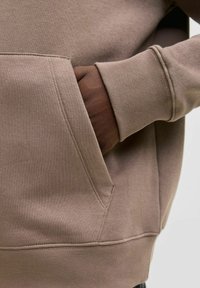 Καφέ φούτερ με τσέπη καγκουρό στο μπροστινό μέρος, διαθέτει υφασμάτινο περίγραμμα και ριγέ μανσέτες. Λείο ύφασμα με γήινο χρώμα.