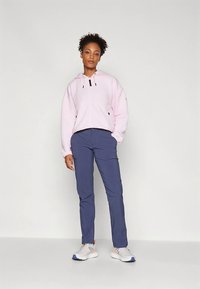 Světle růžová mikina na zip s kapucí a texturovanou látkou, v kombinaci s tmavě modrými kalhotami a bílými sportovními botami s oranžovými detaily.