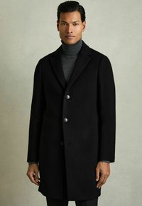 Sort uldfrakke med spids krave, tre knapper og sidelommer. Bæres over en grå turtleneck, har en skræddersyet pasform.