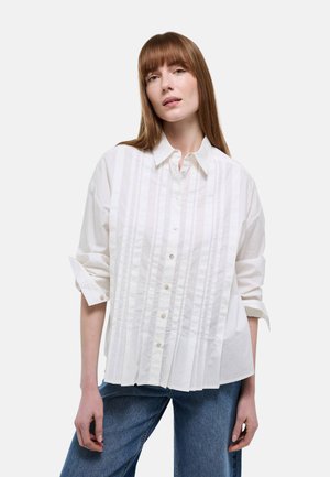 Femme aux cheveux raides rouges portant une chemise blanche ample à boutons avec des plis verticaux et un jean bleu, debout devant un fond uni.