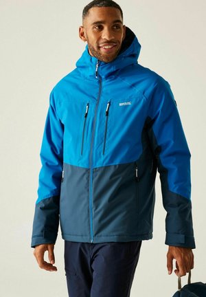 Homme souriant, portant une veste d'extérieur à capuche bleue et marine, tenant une sangle de sac, sur un fond clair uni.